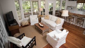 Una sala de estar con un sofá blanco, una mesa de centro de madera y un televisor. Amplias ventanas ofrecen vistas al verdor exterior.