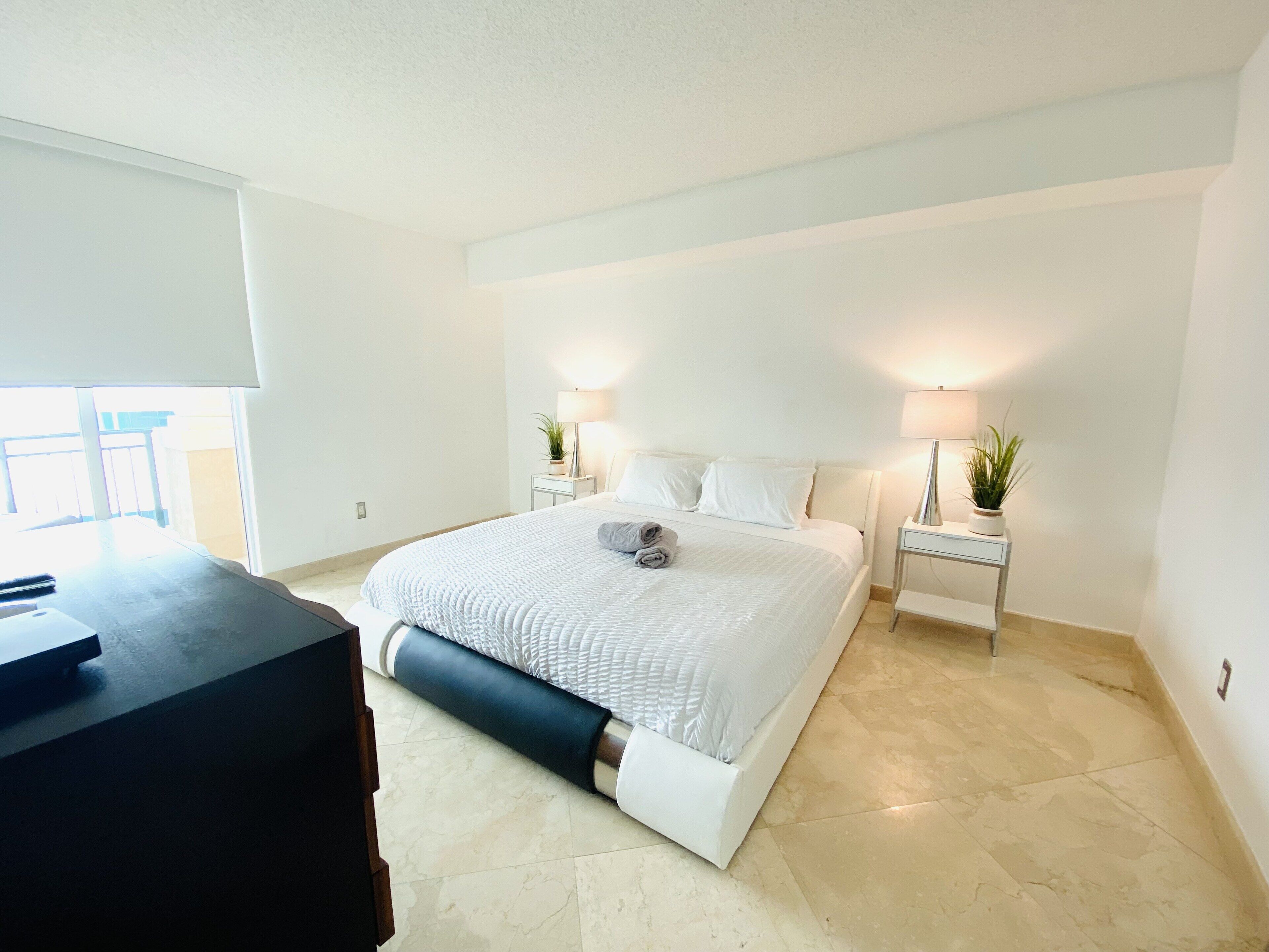 Un dormitorio moderno con una cama grande, dos mesitas de noche con lámparas, un escritorio y vistas al exterior.