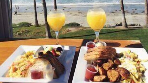 A table with breakfast plates and two glasses of orange juice, overlooking a beach with palm trees and the ocean.