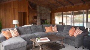 A cozy living room with a gray sectional sofa, wooden beams, a stone fireplace, and large windows.