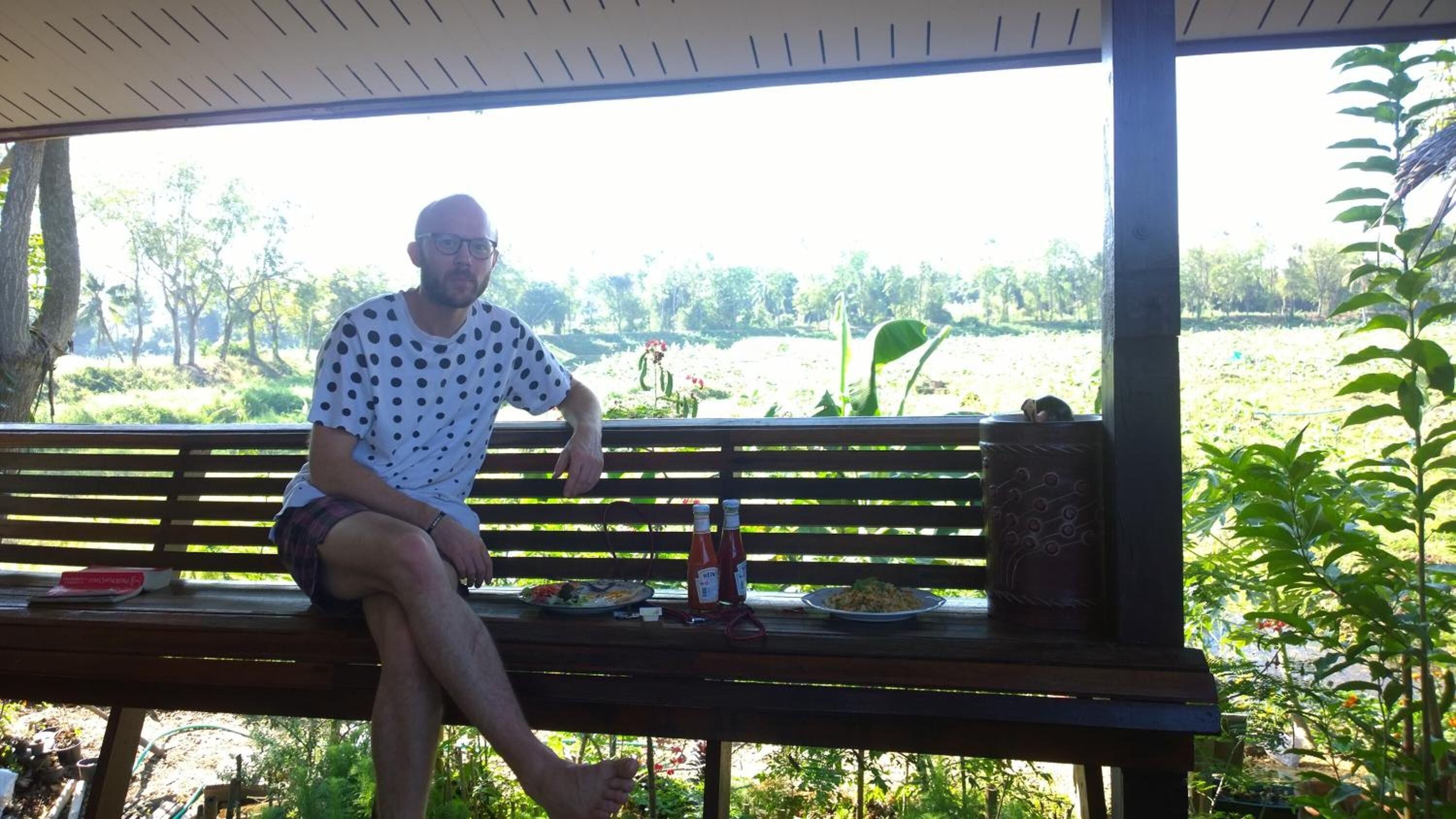 A man sitting on a wooden bench with a table in front of him, outdoors with greenery and trees in the background.