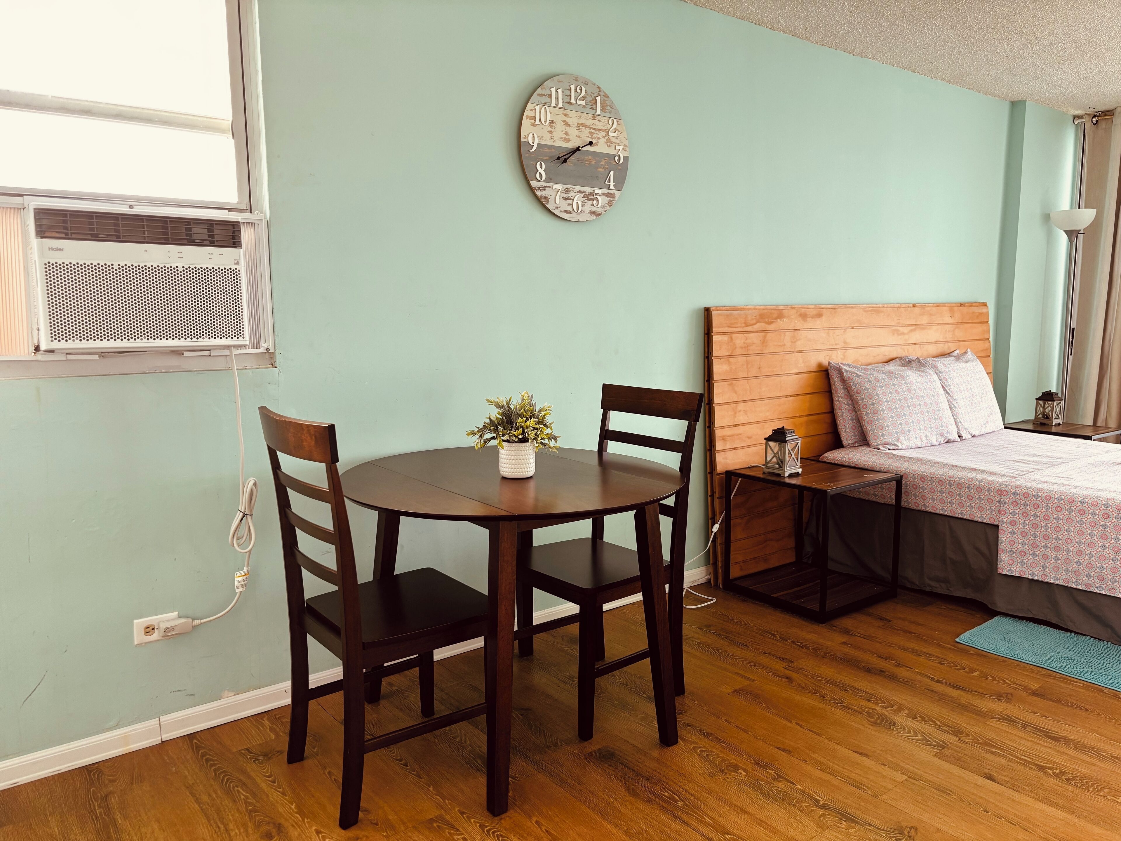 Ein gemütliches Zimmer mit Holztisch und -stühlen, einem Bett mit Holz-Kopfstück und einer Wanduhr.