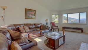 A living room with a brown leather sectional sofa, a wooden coffee table, a chair, and a bench. There is a painting on the wall and a window with a view of mountains. - Amber Sunset - Views! Spacious, Light Bright, Gorge-ous Views! (Mosier)