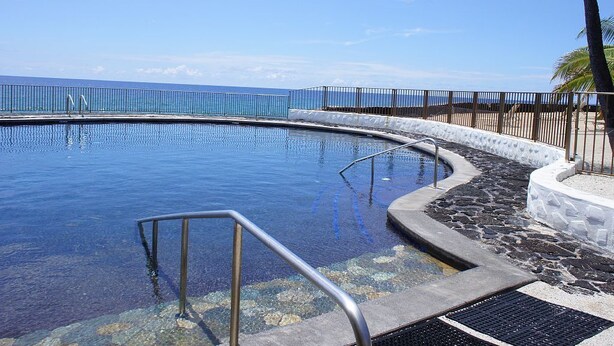 En swimmingpool med metalgel ænder, omgivet af en stenvæg og et hegn, med kig til havet.
