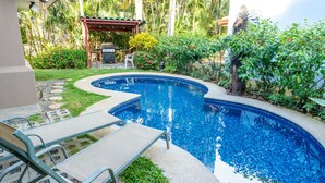 A swimming pool with lounge chairs, surrounded by lush greenery and a covered patio area.