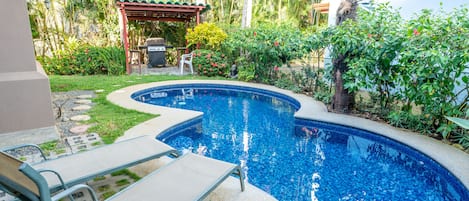 A swimming pool with lounge chairs, surrounded by lush greenery and a covered patio area.