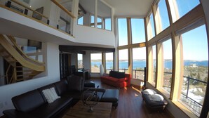 En rummelig opholdsstue med trægulv, en sort lædersofa, en rød sofa og et træsofabord. Store vinduer giver udsigt over havet og et fjerntliggende landskab.