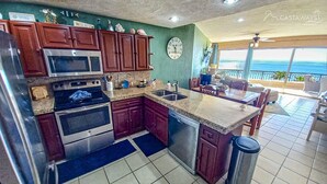 A kitchen with a microwave, oven, and sink. There is a dining area with a table and chairs. A clock is on the wall.