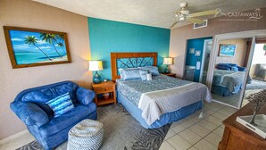 A bedroom with a bed, a blue armchair, a nightstand, a lamp, a painting of a beach, and a ceiling fan.