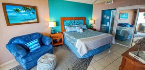 A bedroom with a bed, a blue armchair, a nightstand, a lamp, a painting of a beach, and a ceiling fan. - Sonoran Spa W506 - 2 BR Next to the Pool, High Speed Wi-Fi - Beachfront Upper Floor Luxury Condo (Puerto Peñasco)