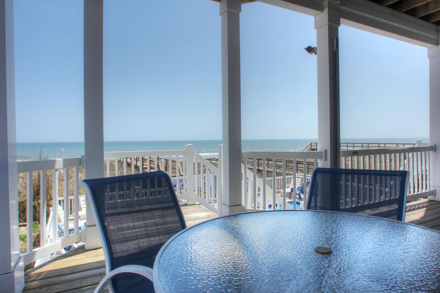 A covered outdoor area with a glass table and two chairs, overlooking a beach with a wooden deck and a clear sky.