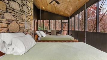 Une chambre avec un mur en pierre, un lit avec une literie blanche, un ventilateur de plafond et de grandes fenêtres donnant sur la forêt.