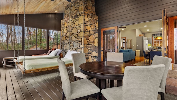 A wooden deck with a stone wall, a ceiling fan, and a dining area with chairs and a table. - Hot tub, Rustic Elegance, In and outside fireplaces, Private, 10 miles to AVL. (Fairview)