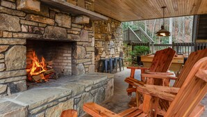 A stone fireplace with a fire, wooden chairs, and a table in a covered outdoor area. - Hot tub, Rustic Elegance, In and outside fireplaces, Private, 10 miles to AVL. (Fairview)