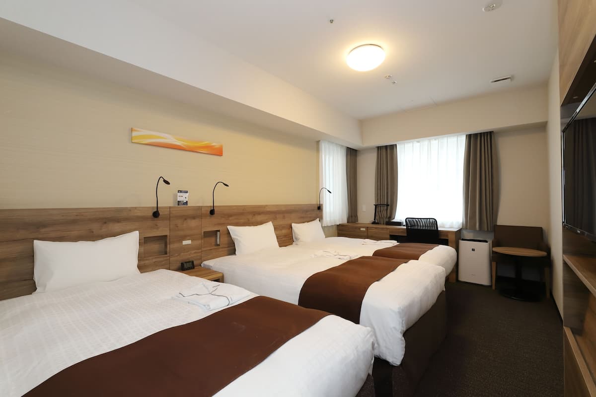 Ett hotellrum med fyra sängar, ett skrivbord, en stol, en tv och ett fönster med gardiner.