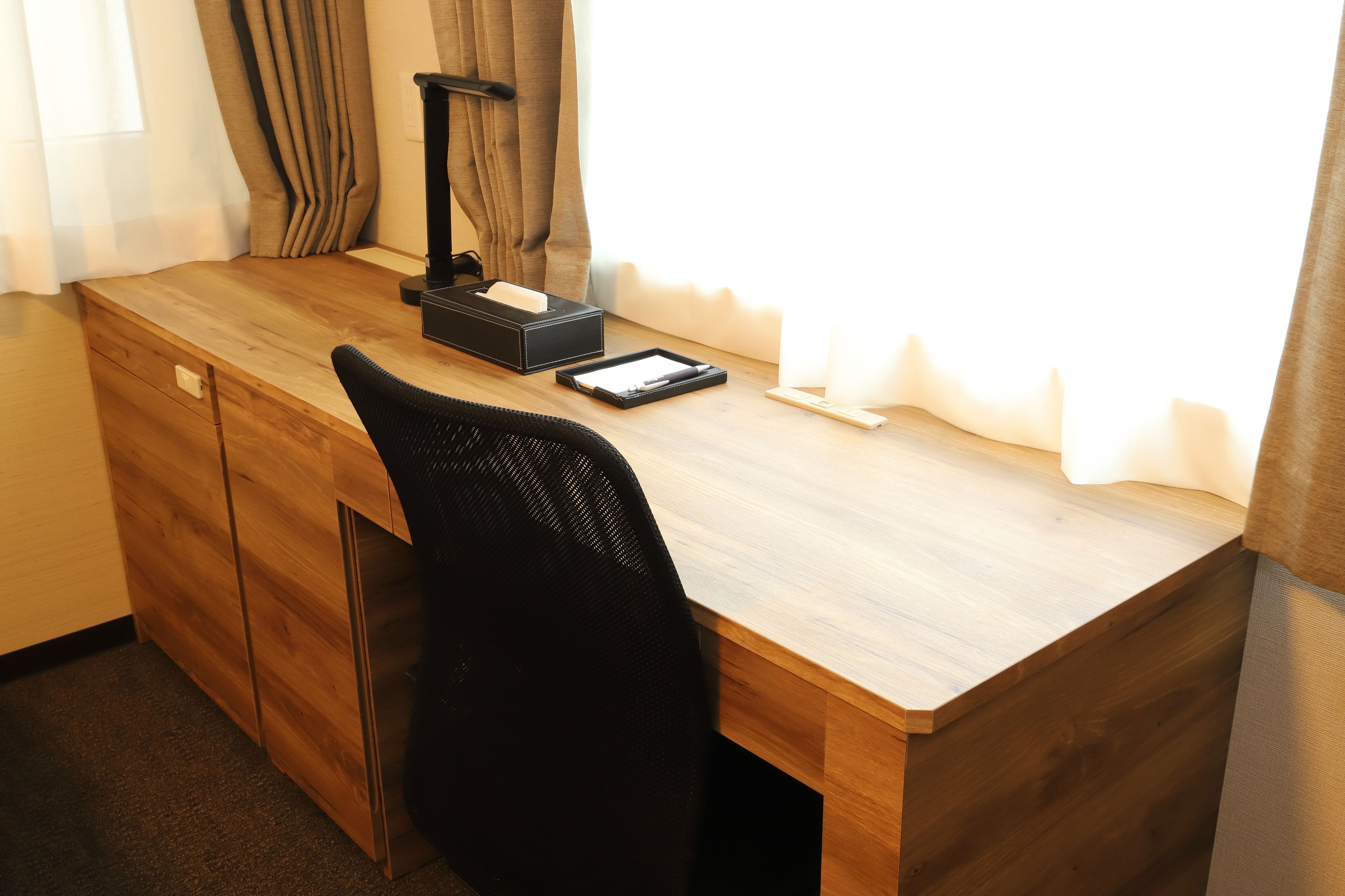 A wooden desk with a black chair, a desk lamp, a tissue box, and a notepad.