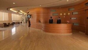 A hotel reception area with a curved wooden reception desk, two receptionists, a sign reading "Cafe Pacifico," and a modern interior design. - Hotel Sewa Grand Faridabad (Faridabad)