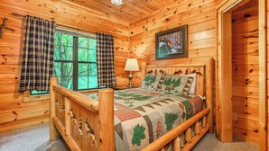 A wooden cabin bedroom with a bed, a window with curtains, a lamp, and a framed picture on the wall. - Making Memories-Theater Room, Pool Table, Arcade, Bear Wood Carvings, Hot Tub (Sevierville)