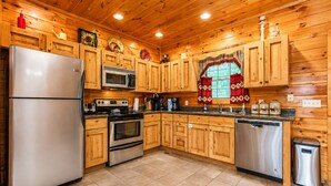 A kitchen with wooden cabinets, stainless steel appliances, a window with red curtains, and a tiled floor. - Making Memories-Theater Room, Pool Table, Arcade, Bear Wood Carvings, Hot Tub (Sevierville)