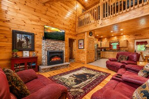 A log cabin interior with a stone fireplace, red leather sofas, a wooden dining area, and a kitchen.
