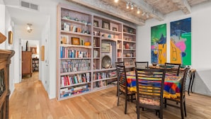 A room with a wooden floor, a bookshelf filled with books and decorative items, a dining table with chairs, and two abstract paintings on the wall.