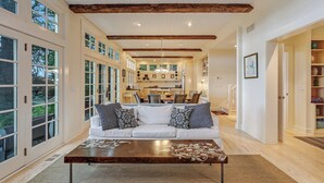 Un vaste espace de vie avec un canapé blanc, des coussins à motifs et une table basse en bois. De grandes fenêtres et une cuisine sont visibles au fond.