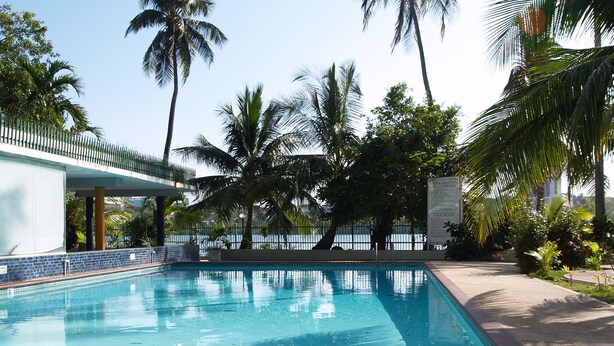 A swimming pool with clear blue water, surrounded by palm trees and a covered walkway. - Beautiful Condo -Ocean View, Best Location, Pool, A Few Steps to All You Want (Santurce)