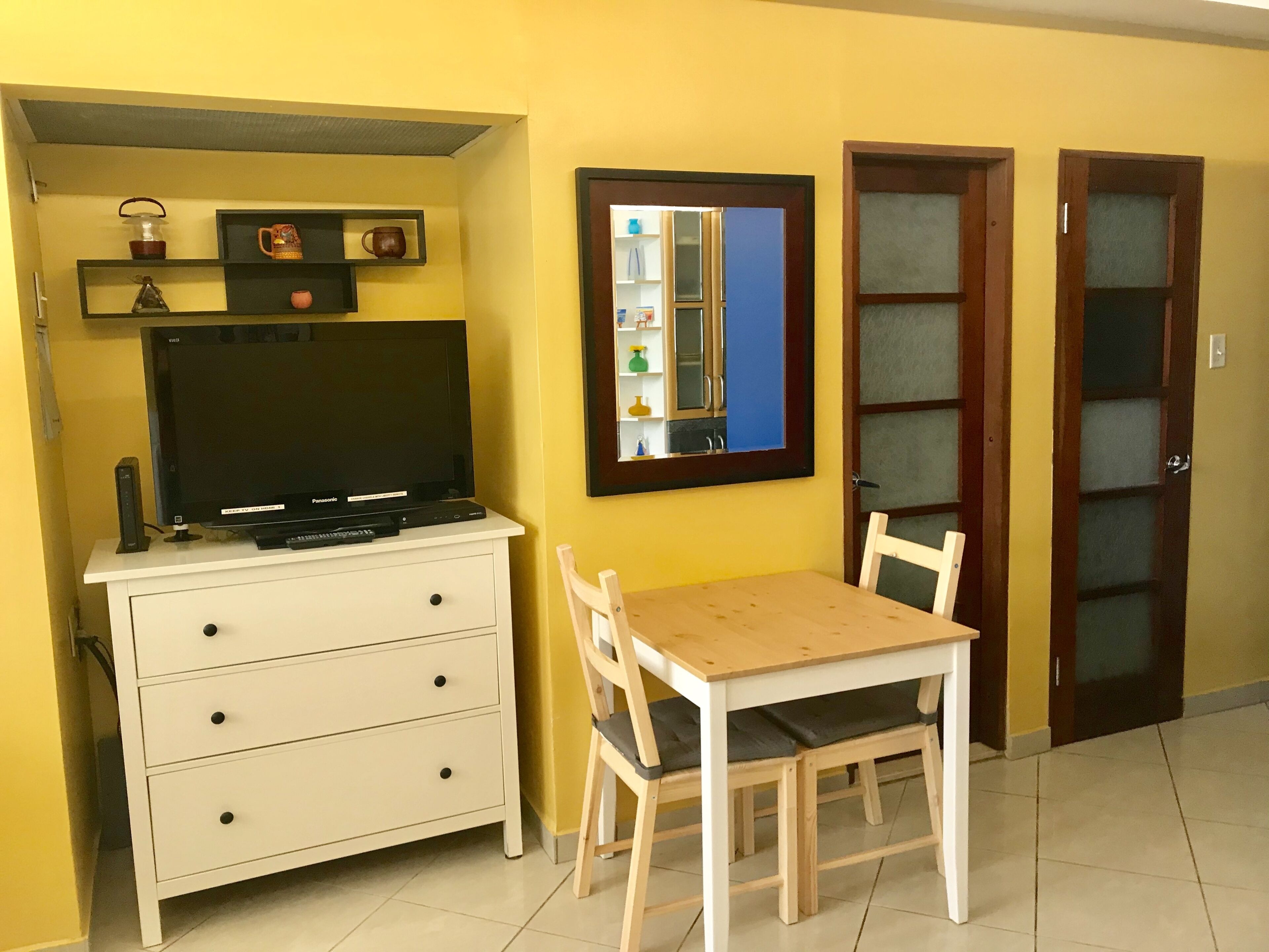 A small room with a TV on a white cabinet, a wooden table with two chairs, and a mirror on the wall.