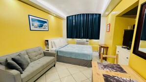 A compact hotel room with a bed, a sofa, a small table, and a TV. - Beautiful Condo -Ocean View, Best Location, Pool, A Few Steps to All You Want (Santurce)