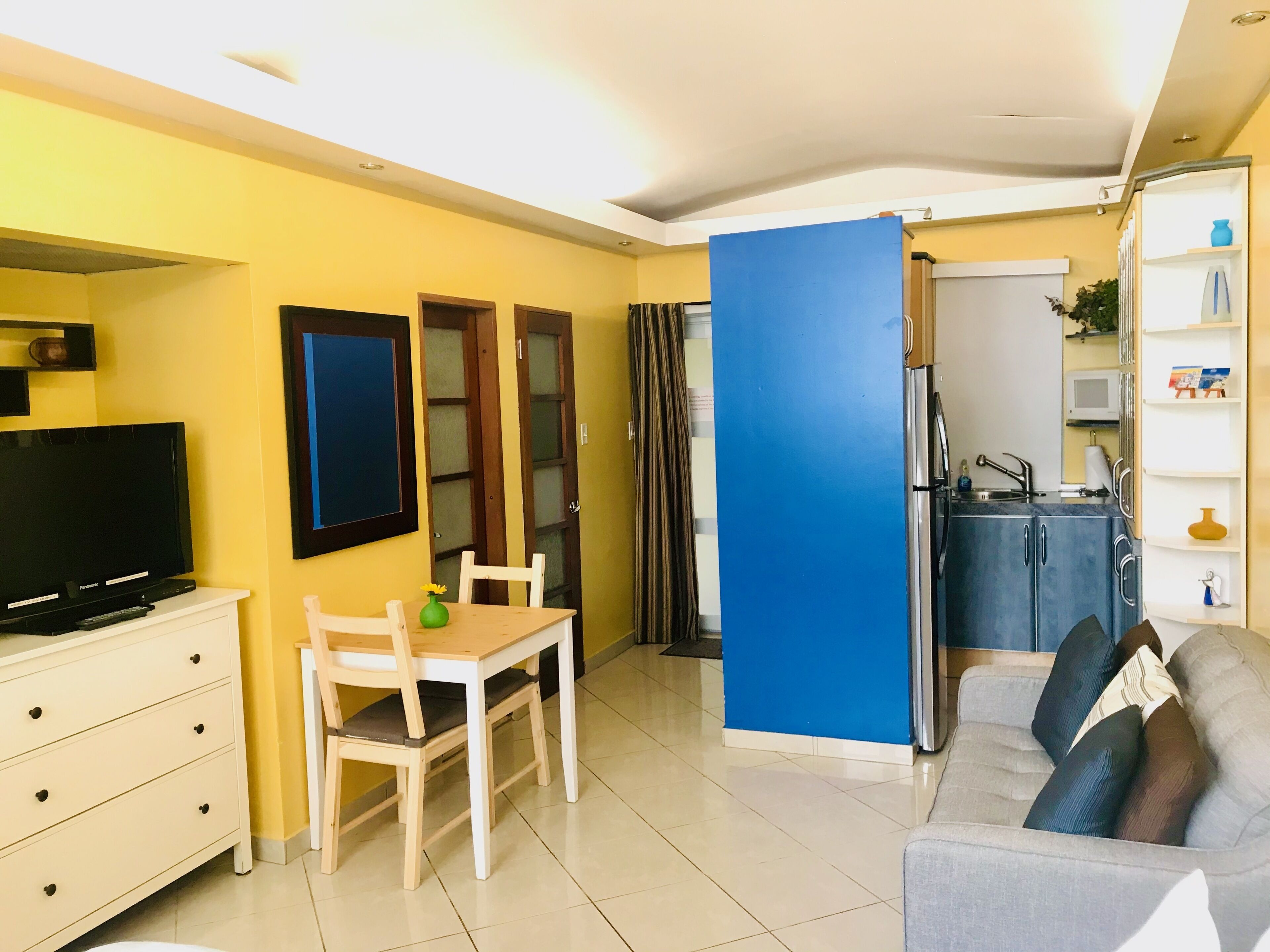 Un ambiente compatto con televisione, un tavolo da pranzo con sedie, un divano, angolo cottura con lavello e una parete divisoria blu.
