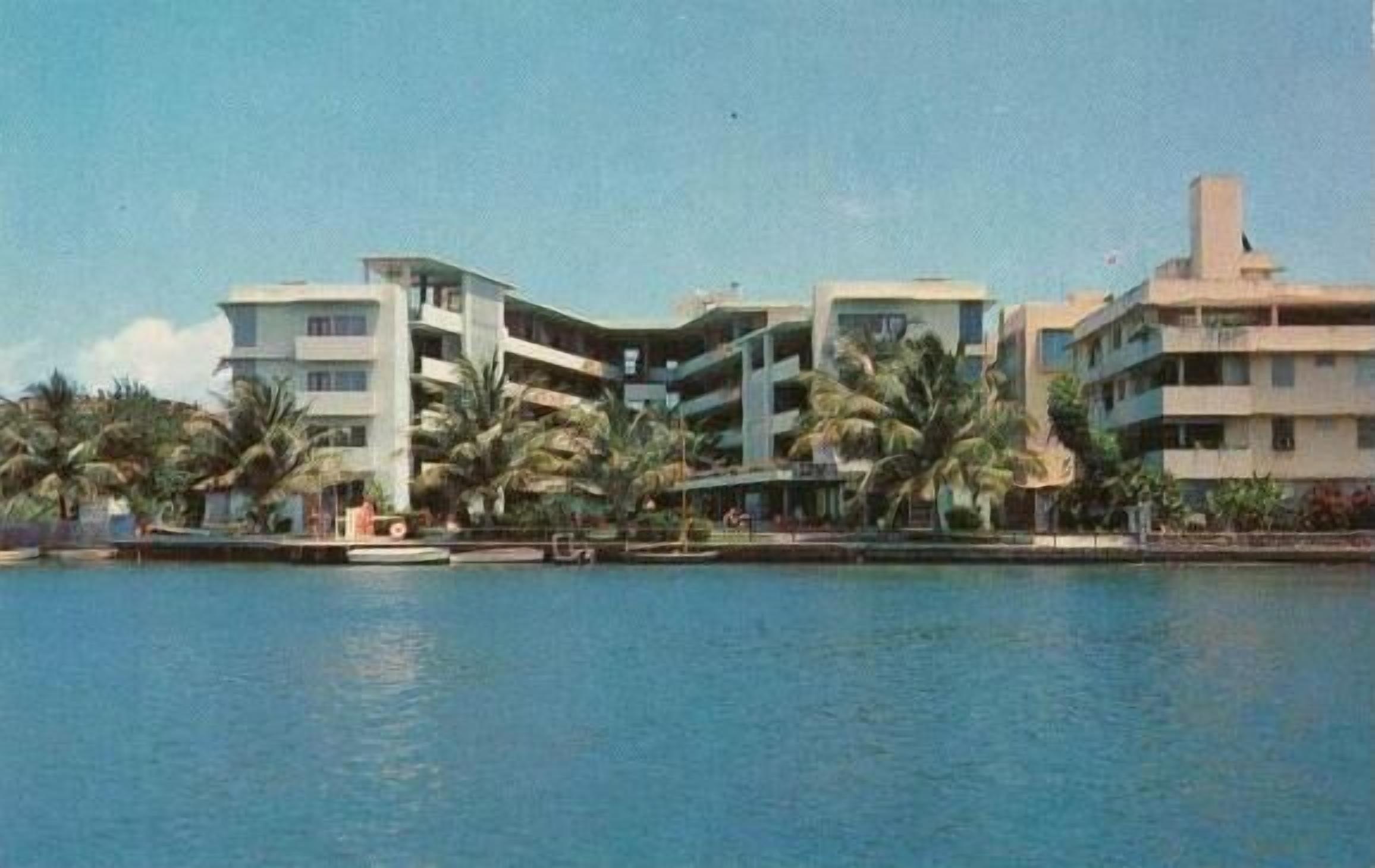 Un hôtel à plusieurs étages doté de balcons, situé près d’une étendue d’eau, avec des palmiers au premier plan.