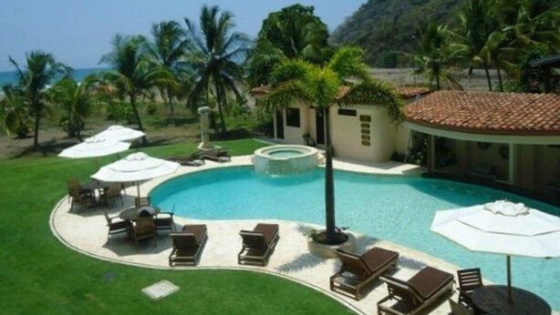A resort pool with lounge chairs and umbrellas, surrounded by palm trees and a view of the ocean.