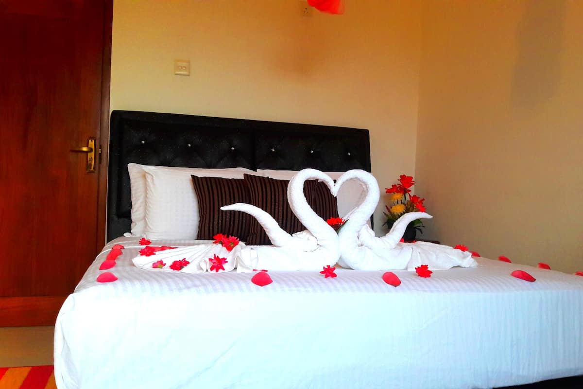 A neatly made bed with a heart-shaped towel arrangement, a vase with flowers, and a wooden door.