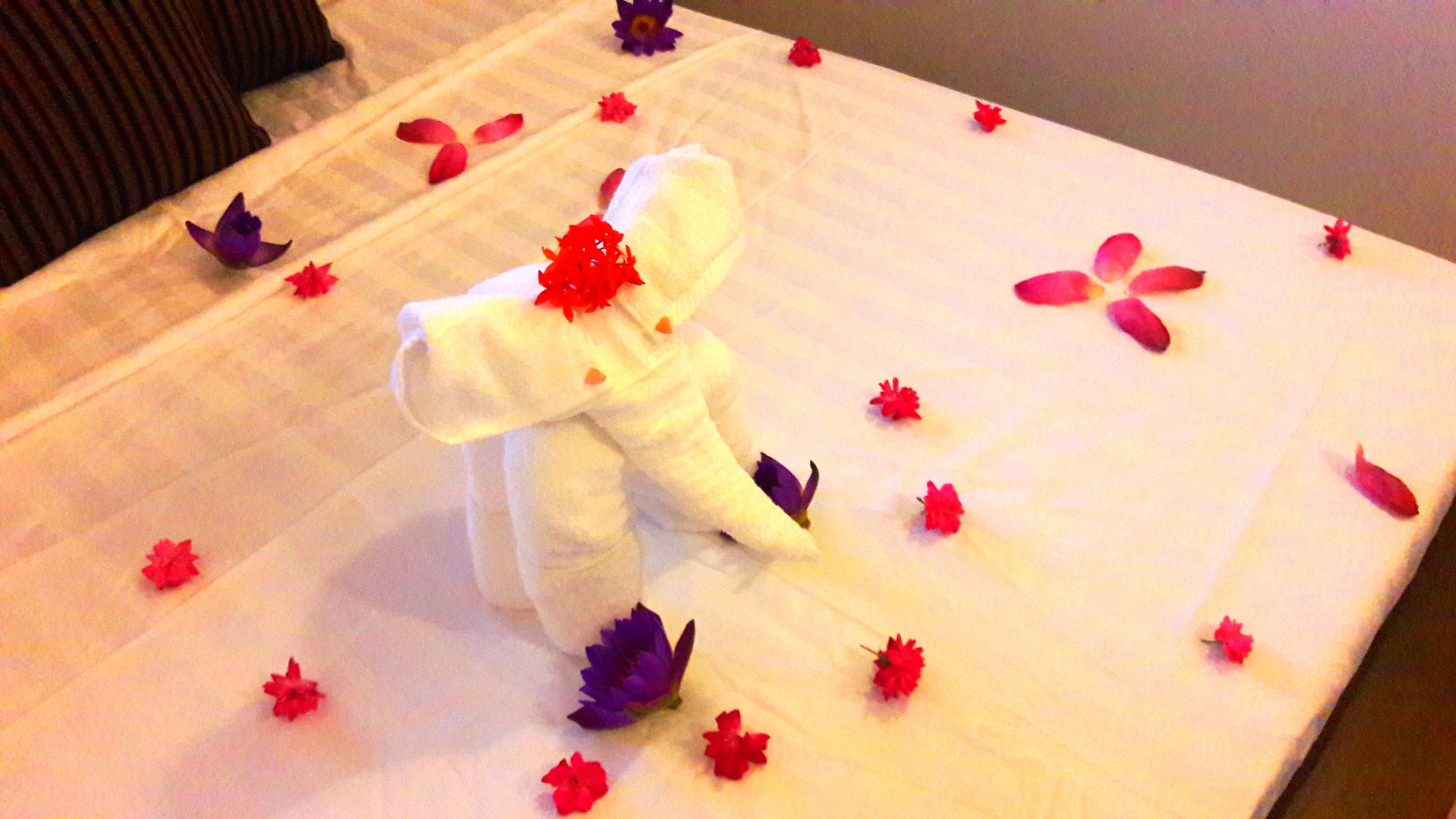 Una mesa ordenada con un mantel blanco, un elefante blanco decorativo y pétalos de flores rojos y morados esparcidos.
