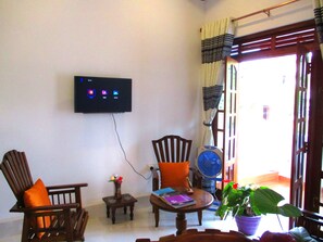 Una sala de estar con un televisor de pantalla plana montado en la pared, dos sillones de madera, una mesita con un libro y un jarrón de flores, y una planta en maceta.