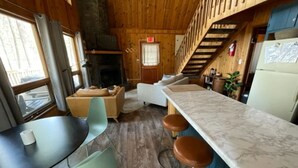 Interior acogedor de una cabaña con techo de madera, chimenea de piedra, zona de comedor con mesa y sillas, barra con taburetes y escalera que sube a un nivel superior.