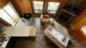 Un acogedor interior de cabaña con piso de madera, un sofá, una mesa de comedor, una chimenea y ventanas grandes.