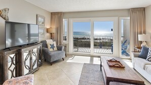 A spacious living room with a large flat-screen TV, comfortable seating, and a balcony with a view of the ocean.