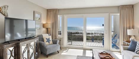 A spacious living room with a large flat-screen TV, comfortable seating, and a balcony with a view of the ocean.