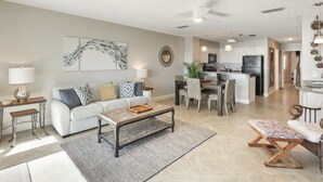 A spacious living area with a sofa, coffee table, and a dining table with chairs. There is a kitchen in the background with cabinets and a refrigerator.