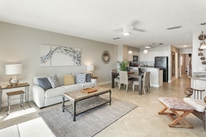 A spacious living area with a sofa, coffee table, and a dining table with chairs. There is a kitchen in the background with cabinets and a refrigerator.