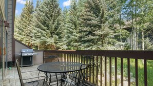 Une terrasse extérieure avec une table ronde et des chaises, un barbecue et une vue sur de grands arbres à feuilles persistantes.