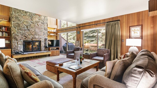 Un salon chaleureux avec une cheminée en pierre, des meubles en bois et de grandes fenêtres donnant sur l’extérieur.