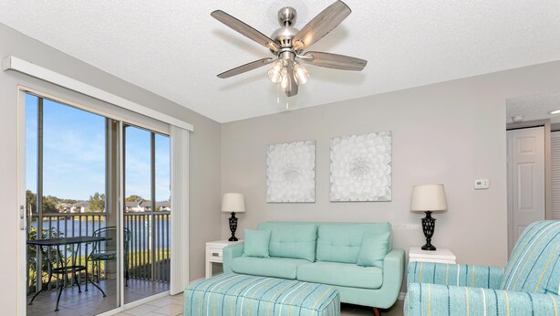A modern living room with a teal sofa, matching ottoman, and armchair, a ceiling fan, and a view of a lake through large sliding glass doors. - Lake Front - Ground Level - 2 bedroom 2 bath (Brandon)