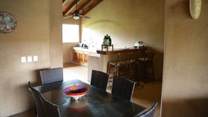 A dining area with a glass table and wicker chairs, a kitchen in the background, and a ceiling fan. - Beautiful serene apartment-saltwater pool, gardens, 200 feet from the beach! (Troncones)