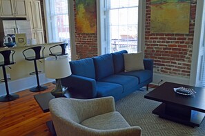 A living room with a blue sofa, a grey armchair, a coffee table, and a lamp. There is a brick wall and a window with blinds. - "Caroline" Julia Street Condo with Balcony (New Orleans)