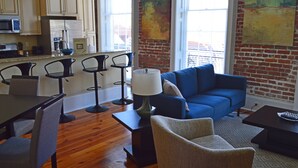 A modern living room with a blue sofa, a dining area with chairs and a table, and a kitchen with wooden cabinets and appliances.