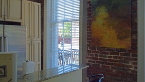A kitchen with white cabinets, a window with blinds, a brick wall, and a painting.
