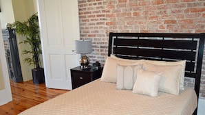 A bedroom with a brick wall, a bed with beige bedding, a nightstand with a lamp, and a potted plant.