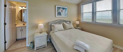 A bedroom with a bed, bedside tables, a nightstand, a window with blinds, a door leading to a bathroom, and a painting on the wall.